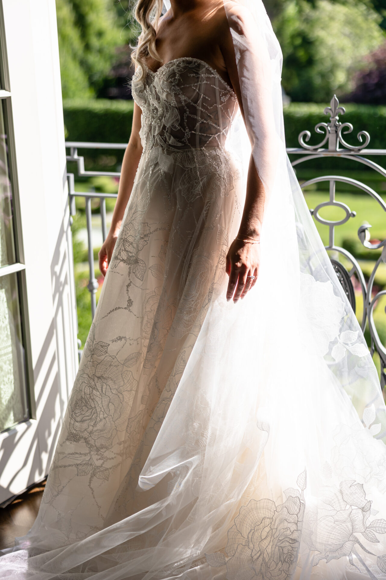 Bride in lace gown by sunlit window, shadowed face.