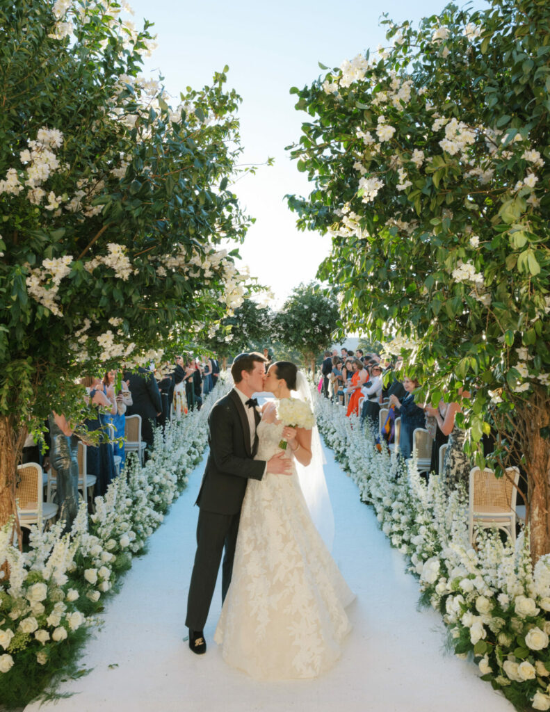 Bride and groom kiss outdoors beneath blooming trees.
