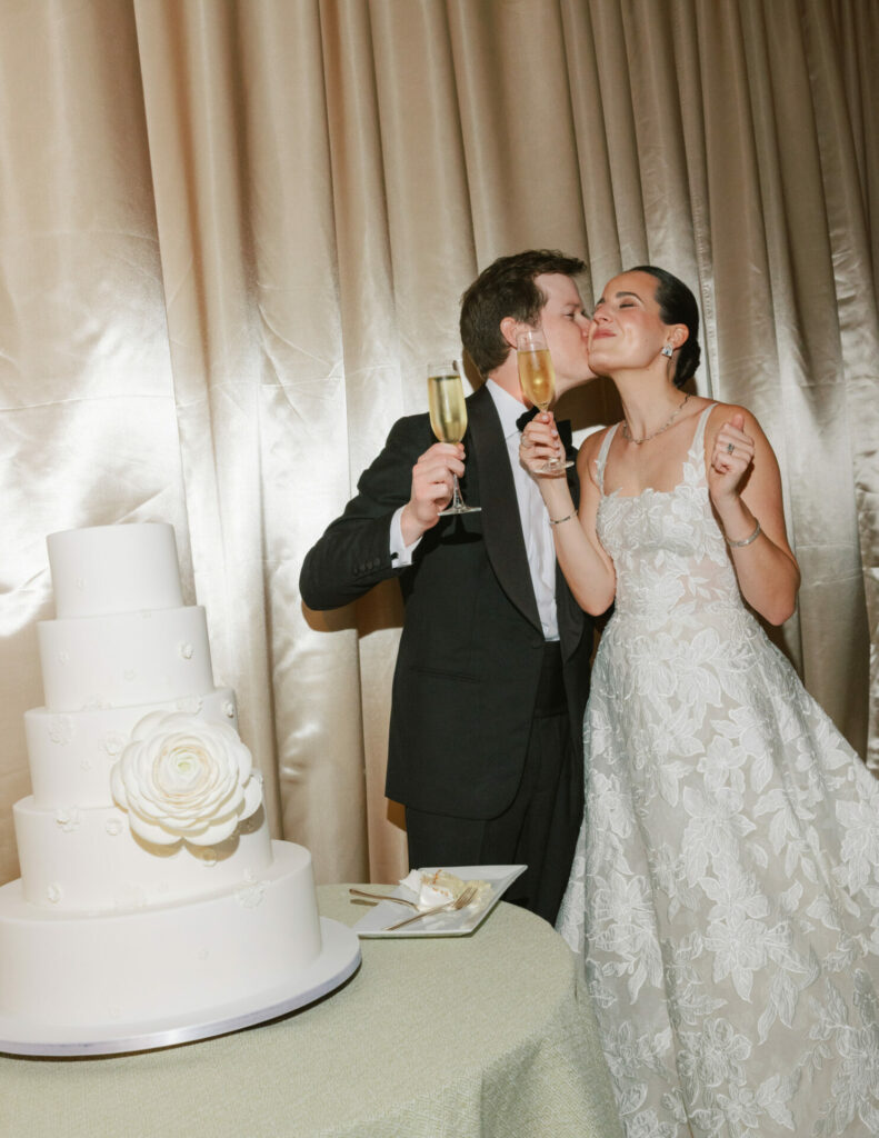 Groom kisses glowing bride by elegant cake, toasting.