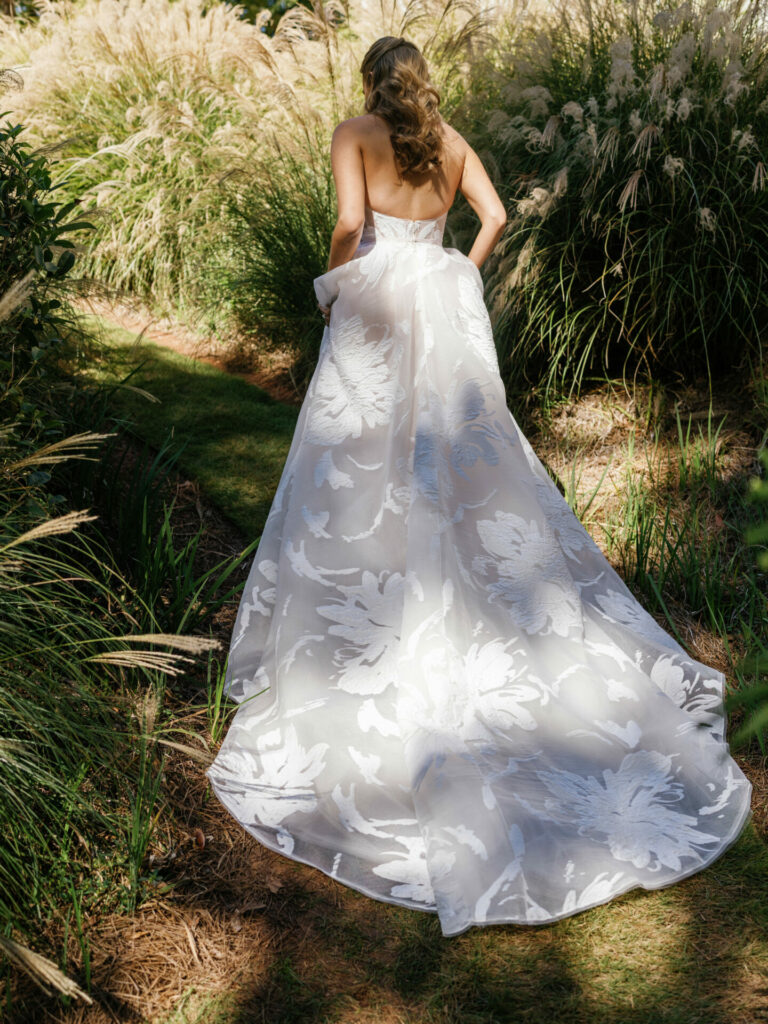 Woman in floral strapless gown walks sunlit grassy path.