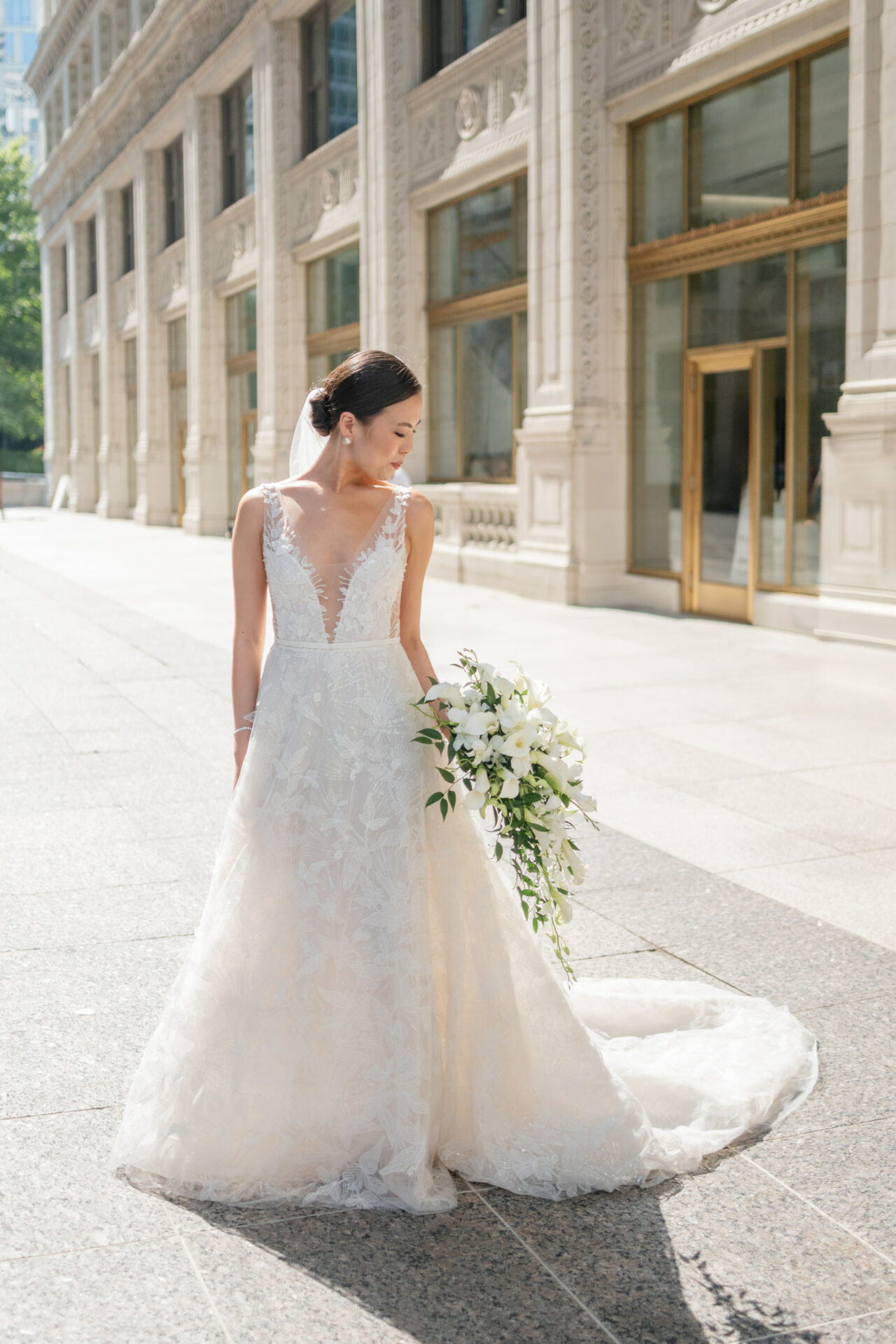 Bride in lace gown with bouquet outside grand building.