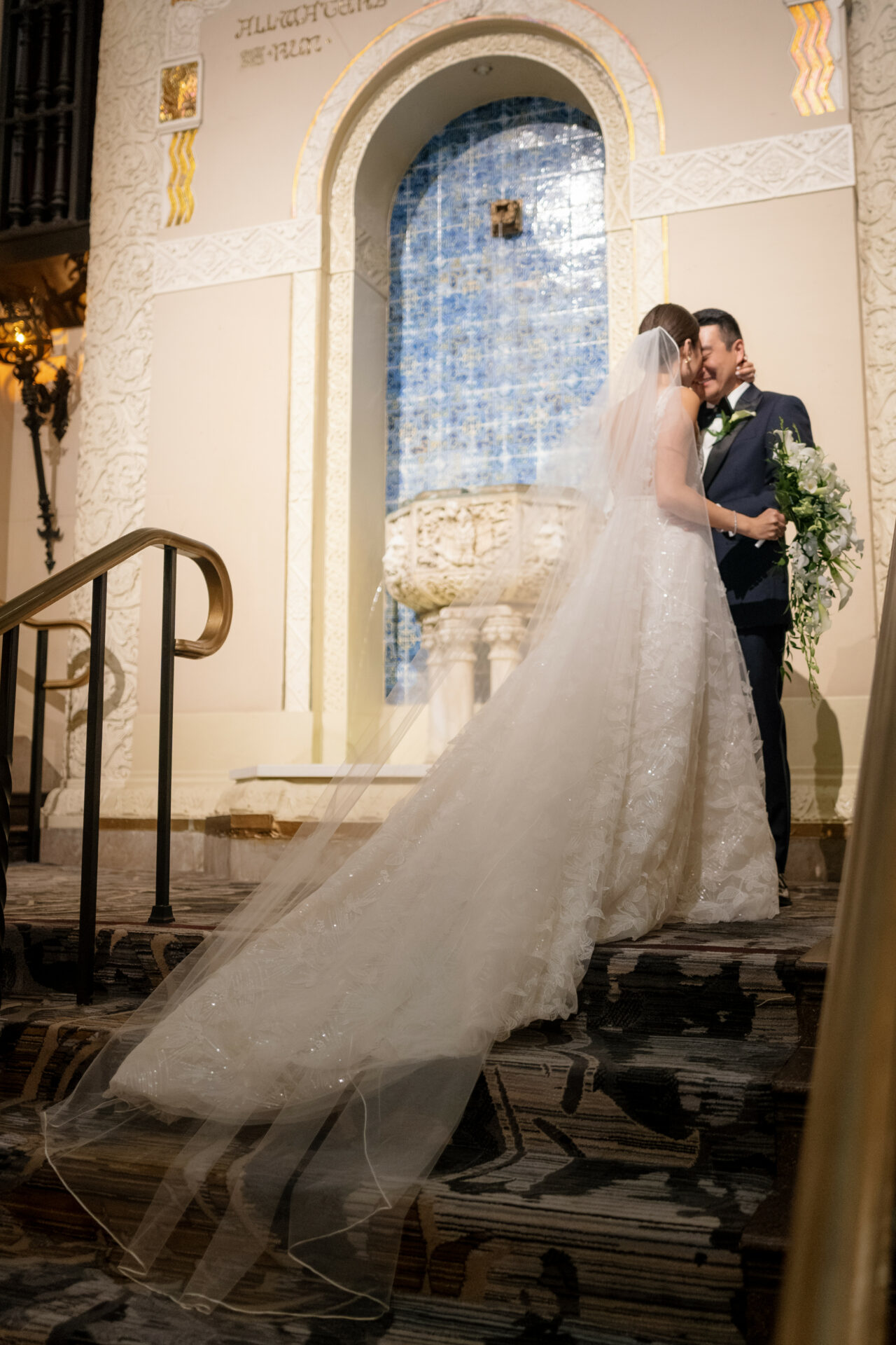 Bride and groom smiling atop staircase, veil flowing.