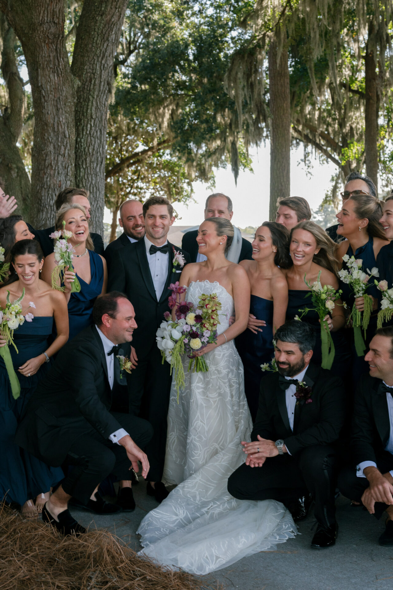 Wedding party smiles outdoors beneath sunlit trees.