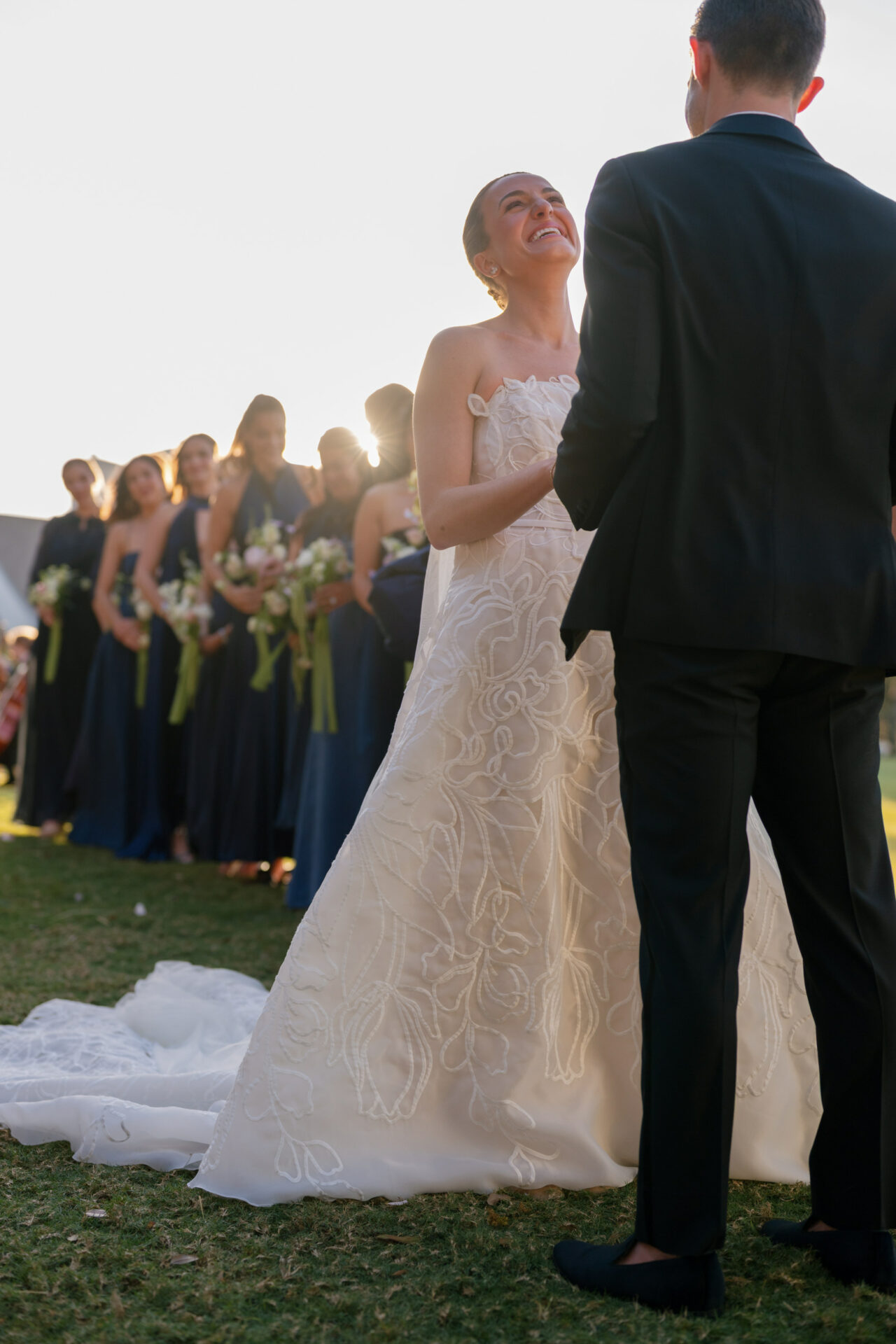 Bride smiling at groom outdoors, bridesmaids and sunlight.