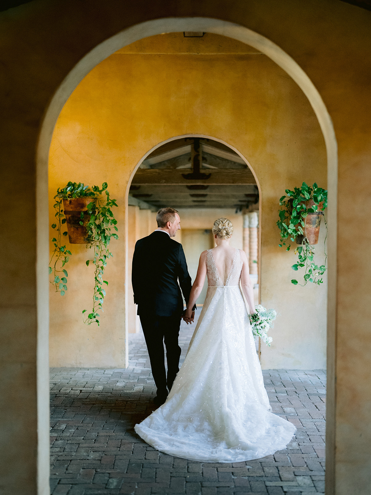 Bride and groom hold hands, walking sunlit corridor.