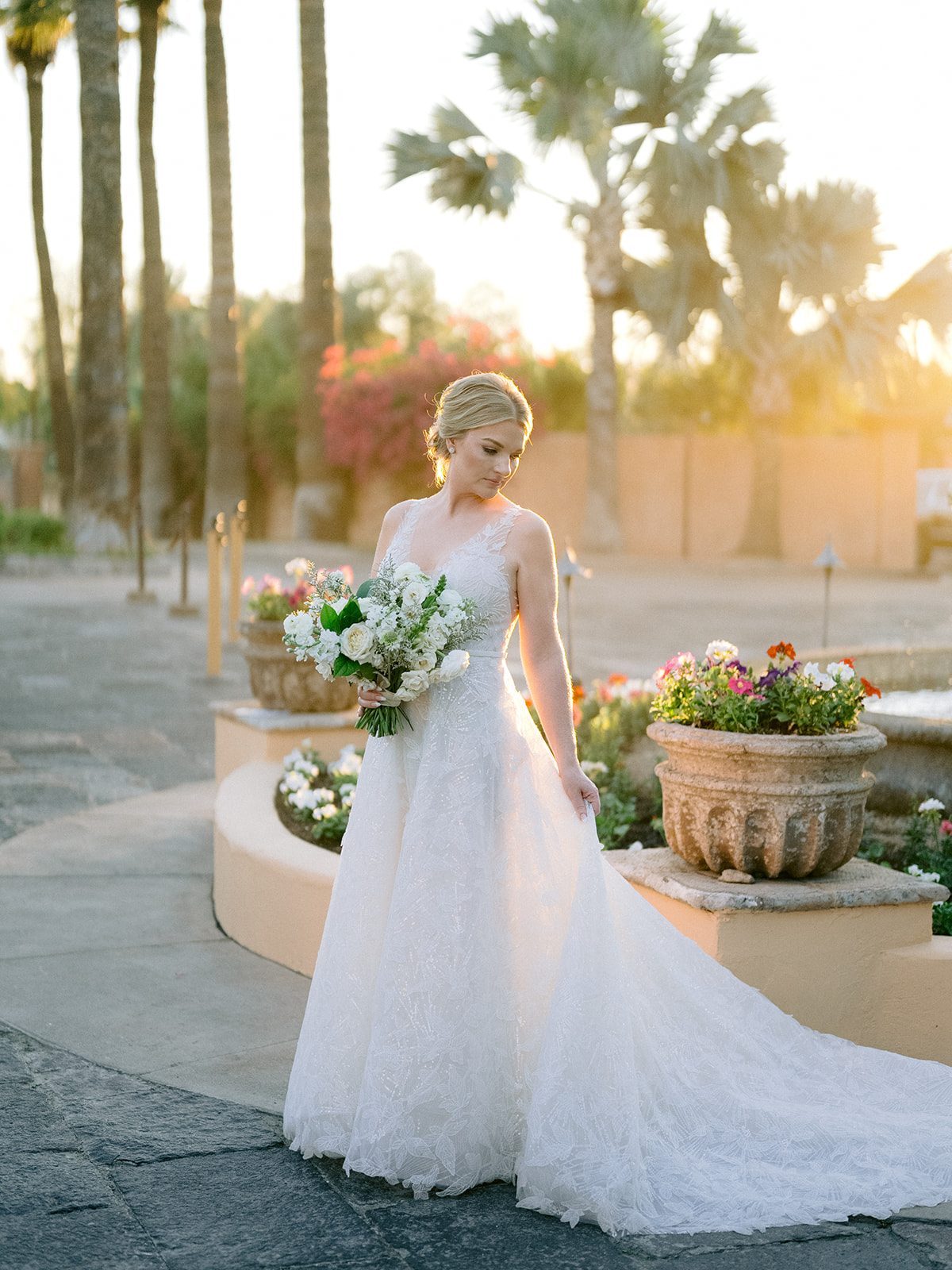 Bride in lace gown holding bouquet, sunny palms behind.