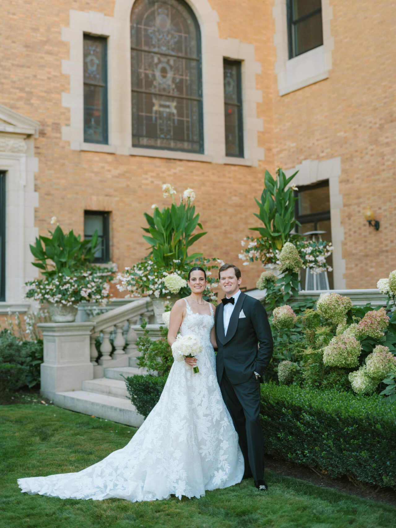 Bride and groom pose, smiling, in luxurious garden.