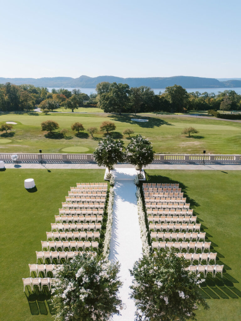 Outdoor wedding with beige chairs, flower-lined aisle, hills.