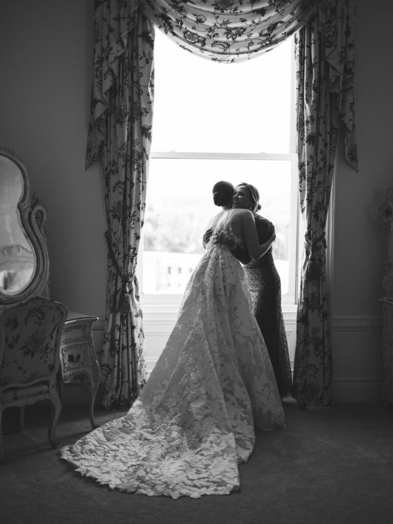 Bride in textured gown hugs woman by ornate window.