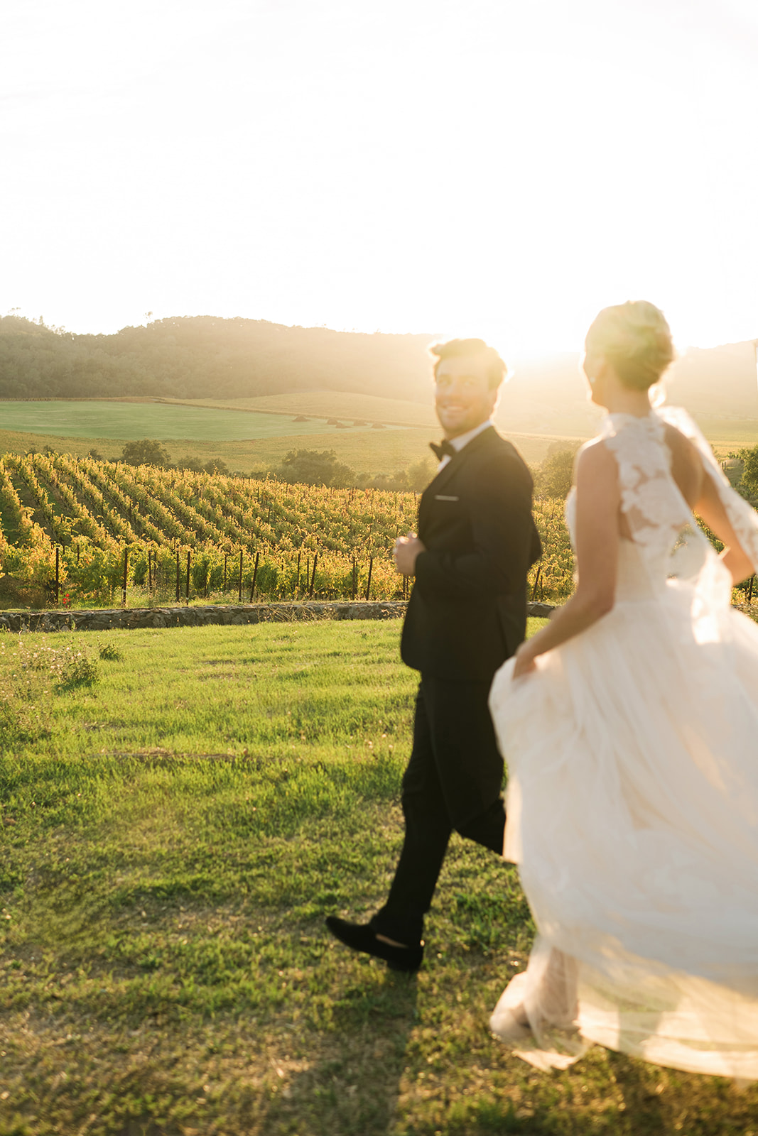 Bride and groom walk in vineyard at sunset, smiling.