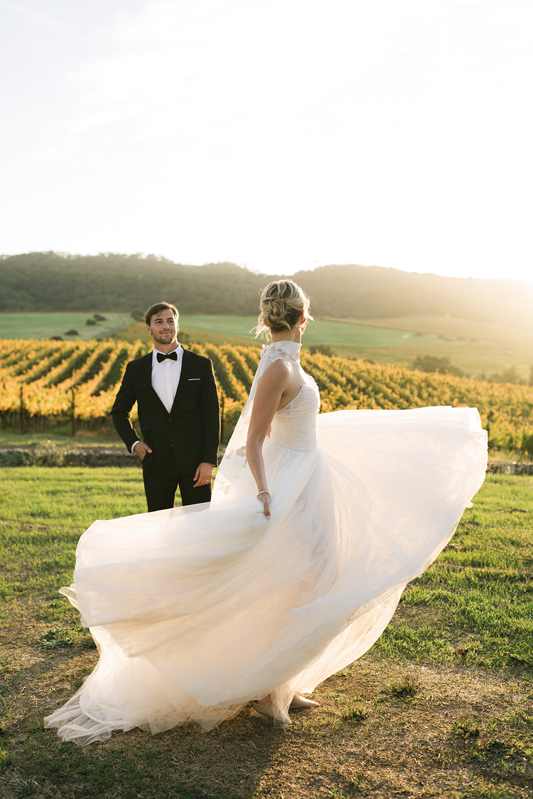 Bride twirls in white dress, groom watches, vineyard backdrop.