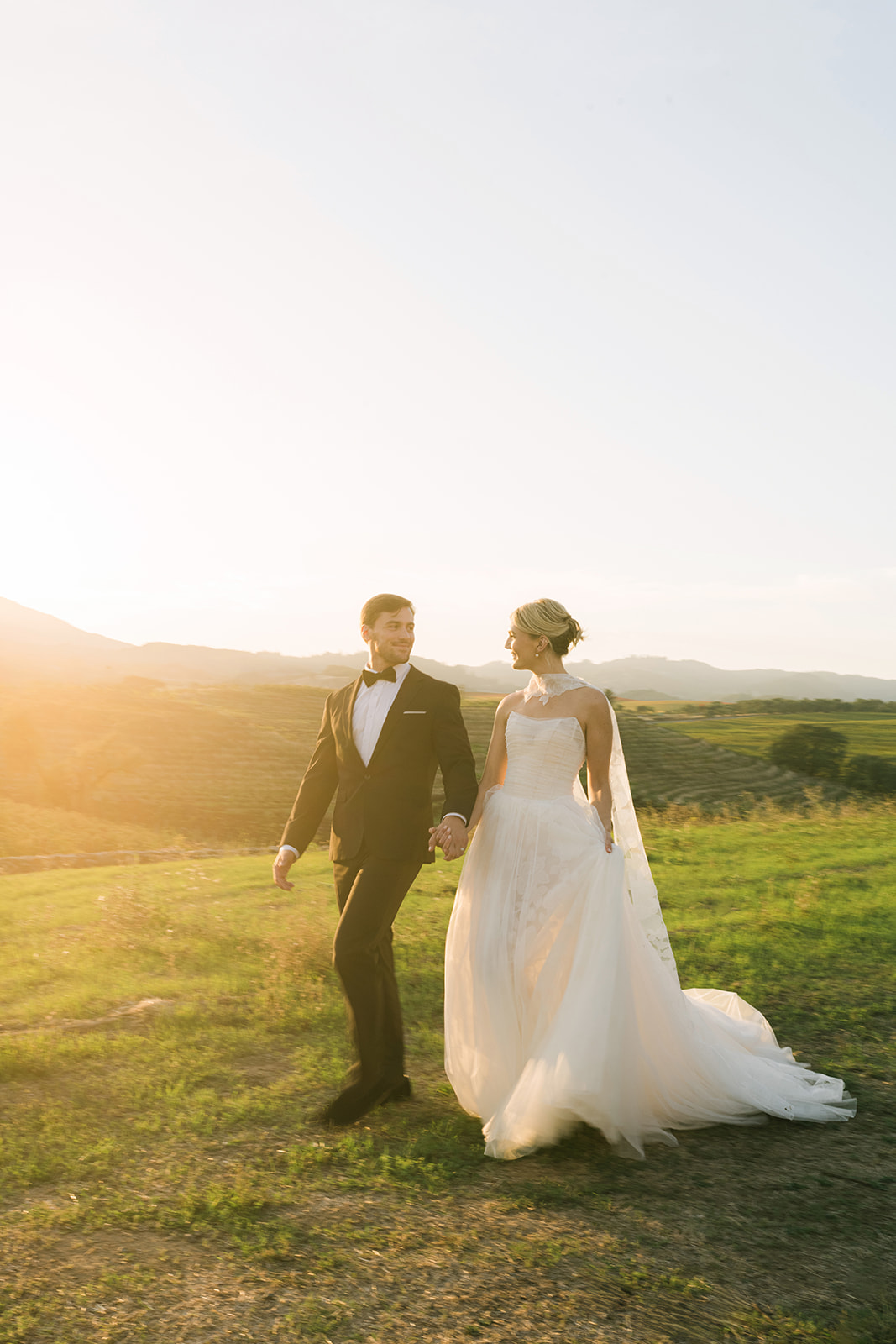 Bride and groom walk at sunset in grassy field.