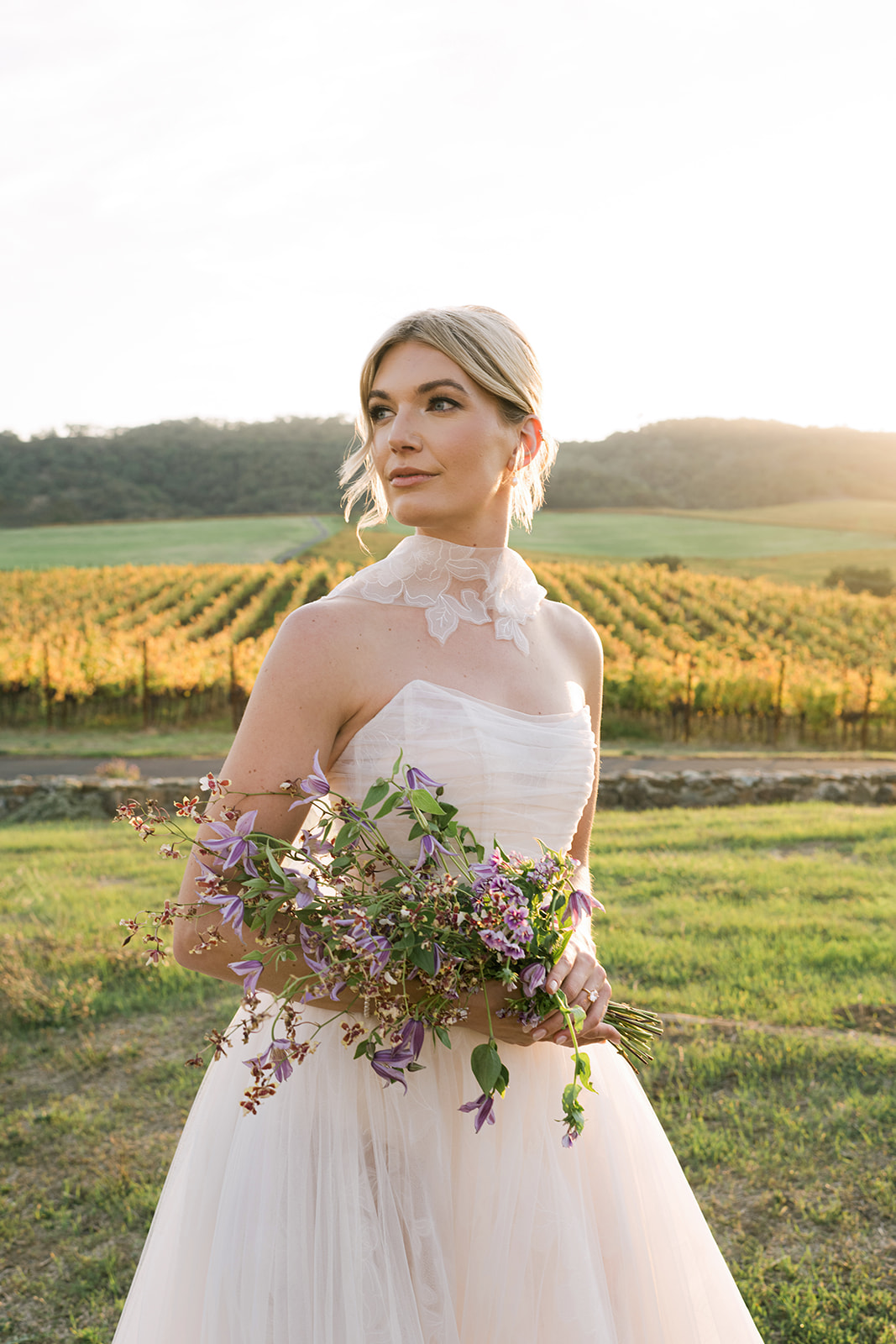 Bride in white dress with purple bouquet outdoors.