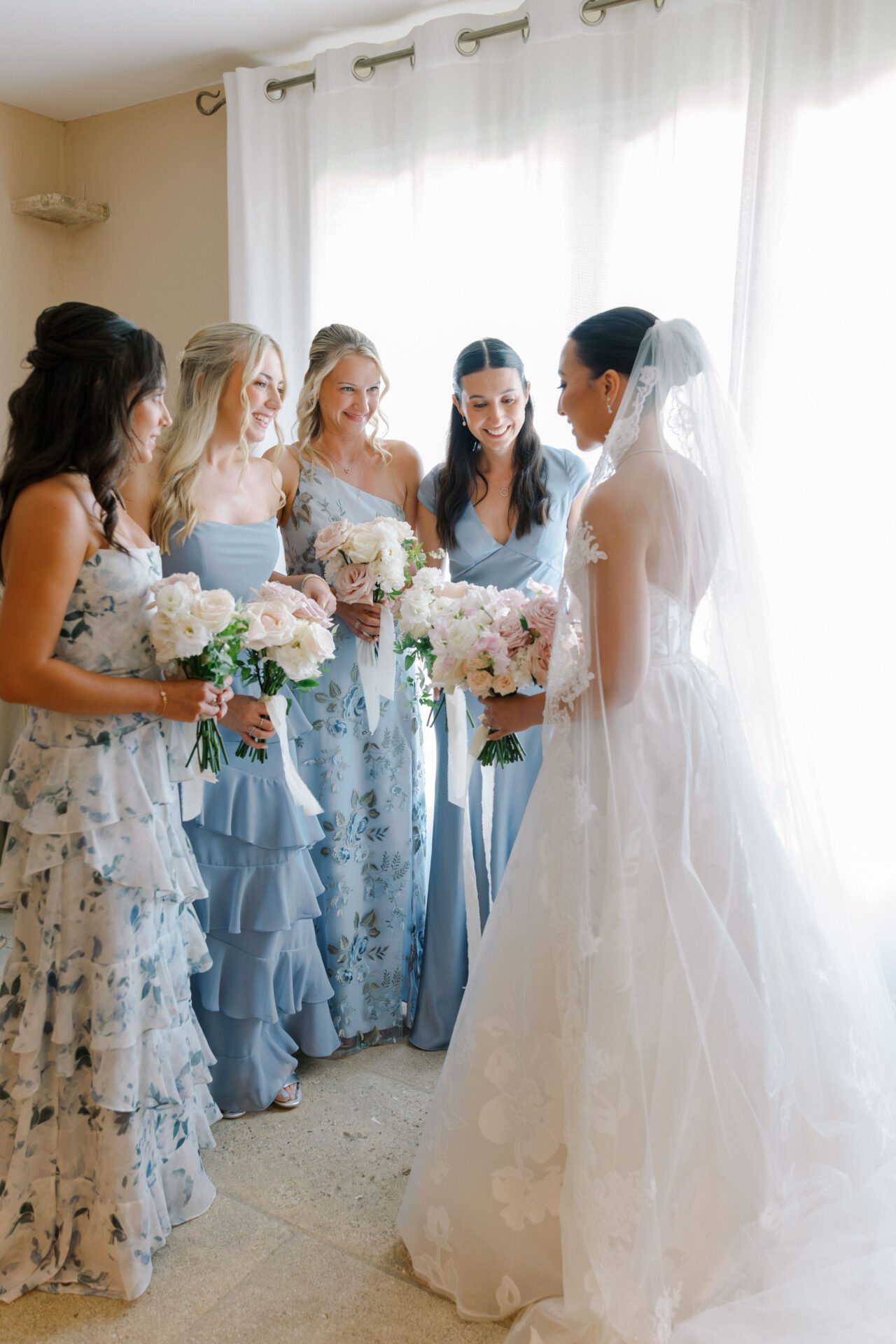Bride with four bridesmaids in sunlit room, smiling.