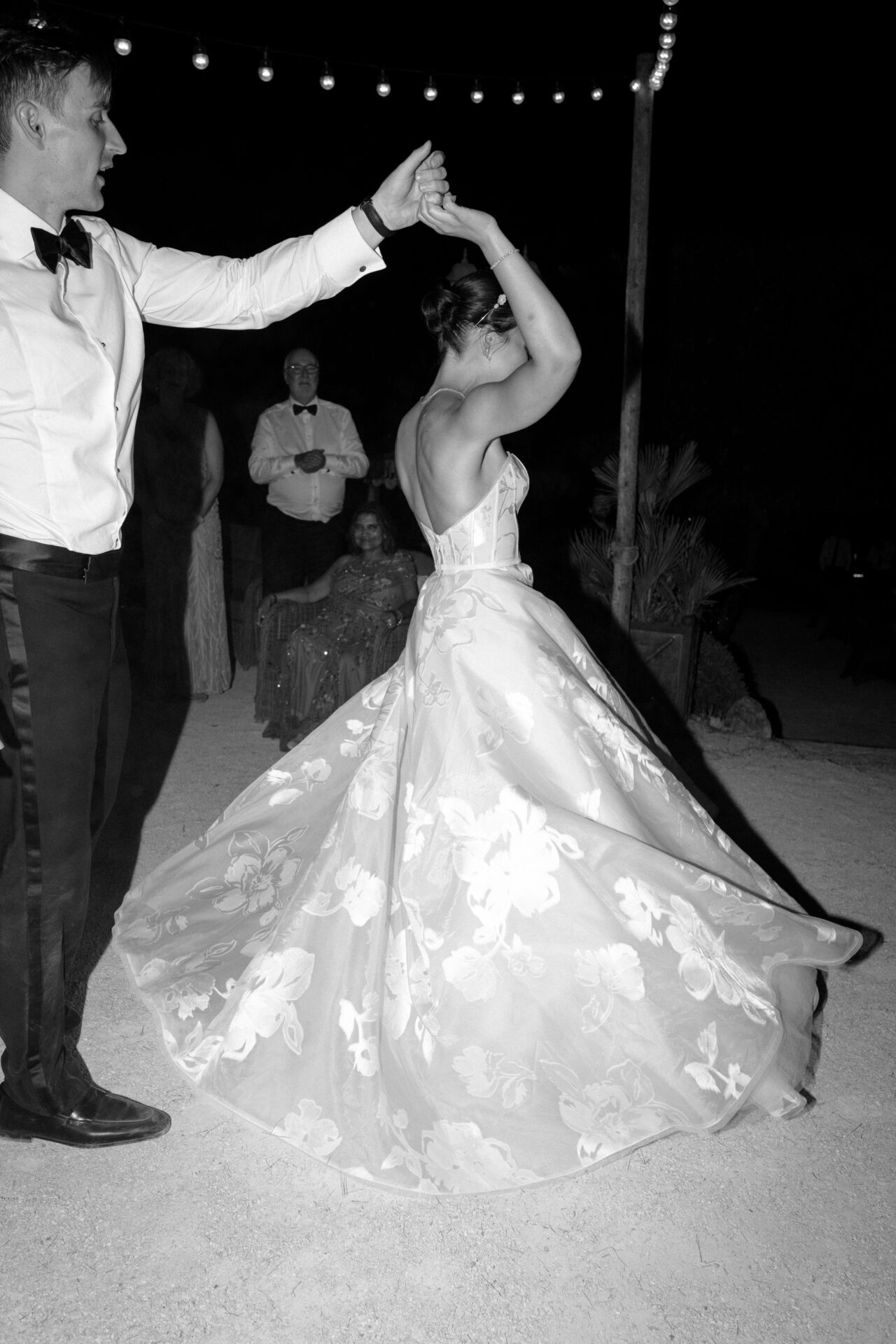 Couple dancing beneath string lights, guests watching.
