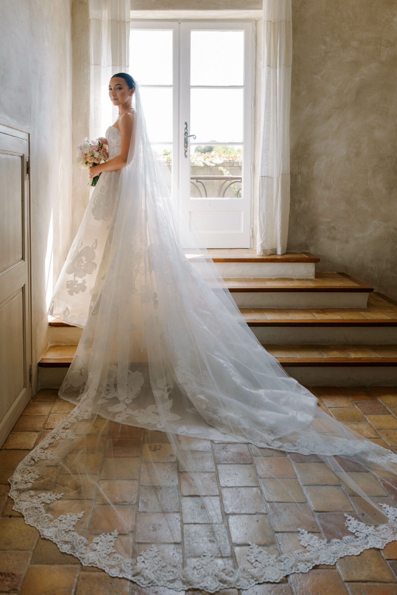 Bride in lace veil on sunlit steps, bouquet in hand.