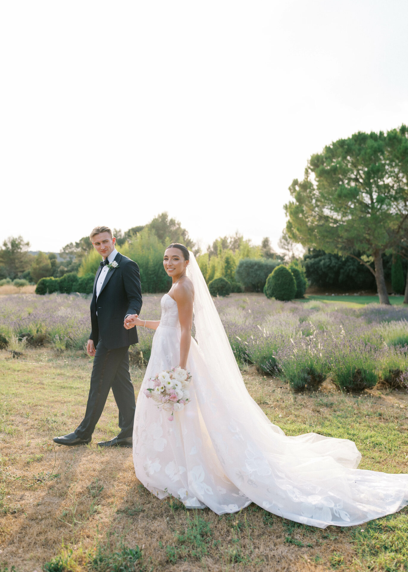 Bride and groom walk hand-in-hand in sunlit garden.