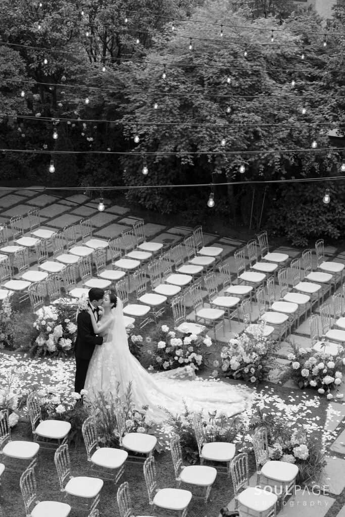 Bride and groom embrace under string lights, black-and-white.