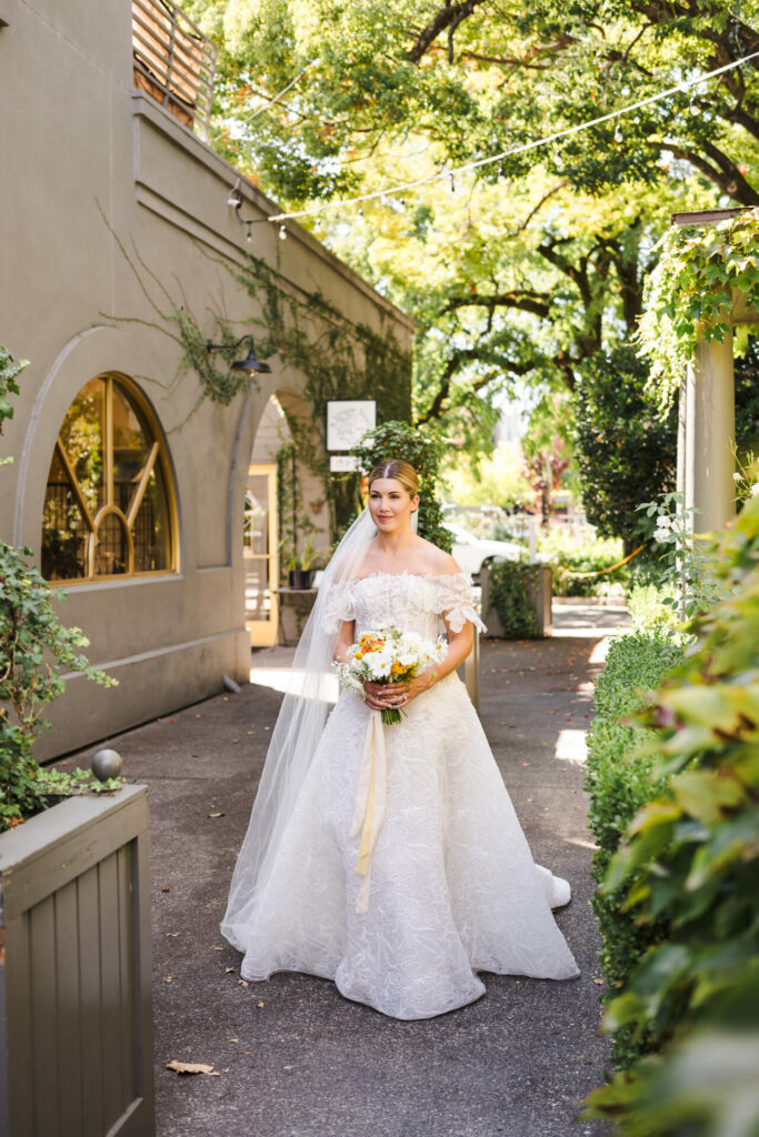 Bride in SS2026 gown holds bouquet outdoors, sunny.
