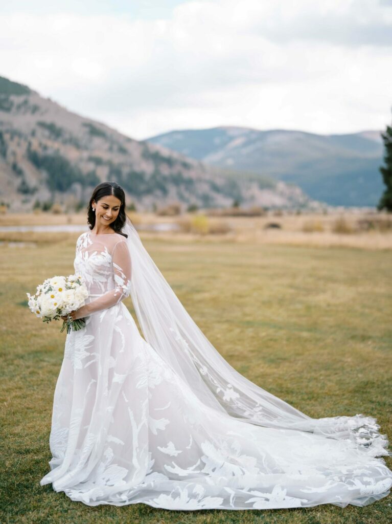 Bride in white lace gown on grassy field, mountains behind.