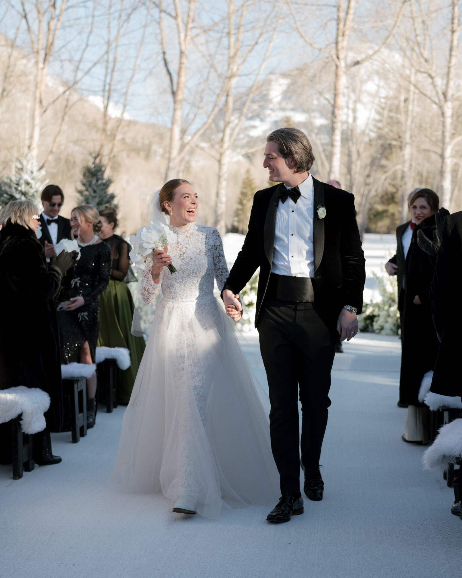 Bride and groom walk outdoor aisle, snowy trees, smiling.