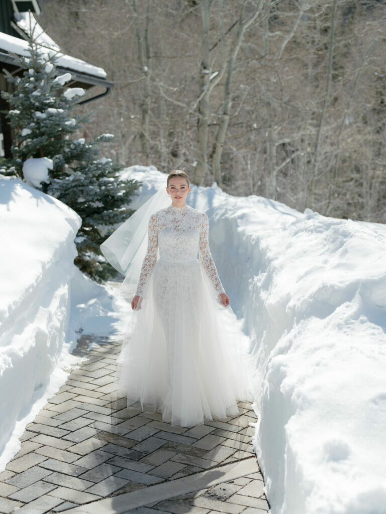 Bride in lace dress on snowy brick path outdoors.