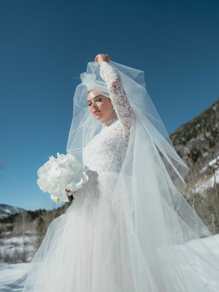 Bride in lace dress holds flowers in snowy mountains.