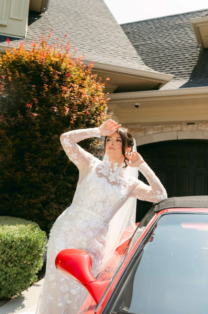 Bride in lace dress poses by red car, shielding sun.