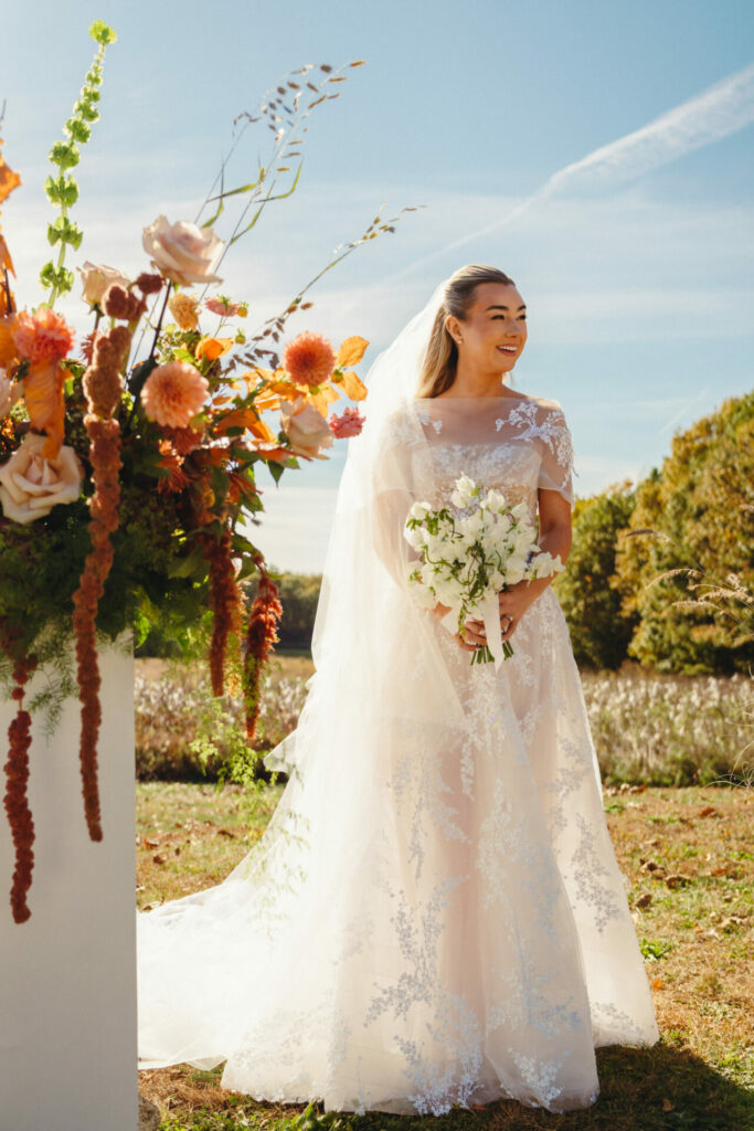 Smiling bride in lace gown, orange florals outdoors.