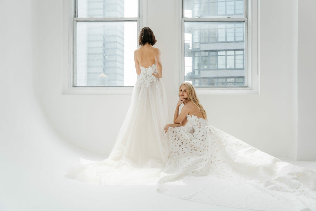 Two women in white bridal gowns pose by windows.