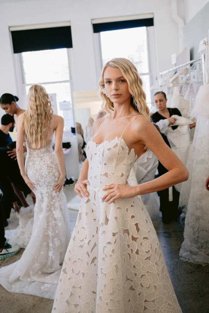 Model in white floral dress backstage at fashion show.