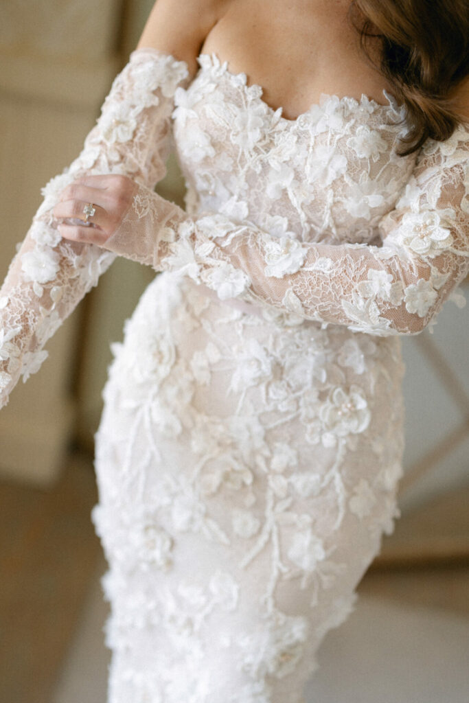 Woman wears lacy off-shoulder wedding dress, ring visible.