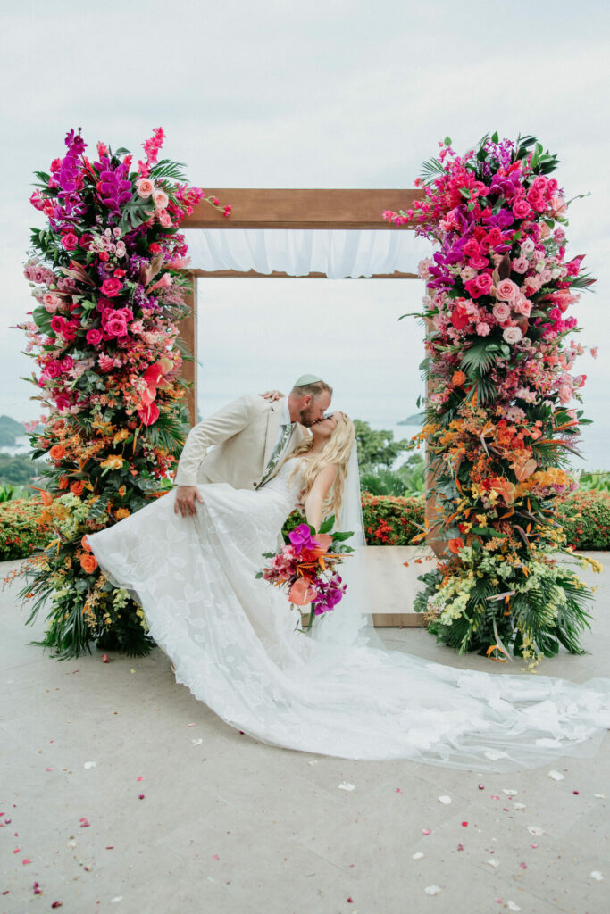 Bride and groom kiss under floral arch, petals around.