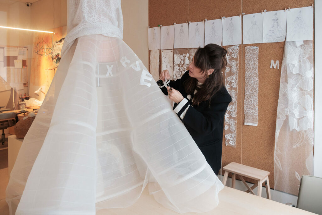 Woman decorates sheer white dress; sketches, lace on wall.