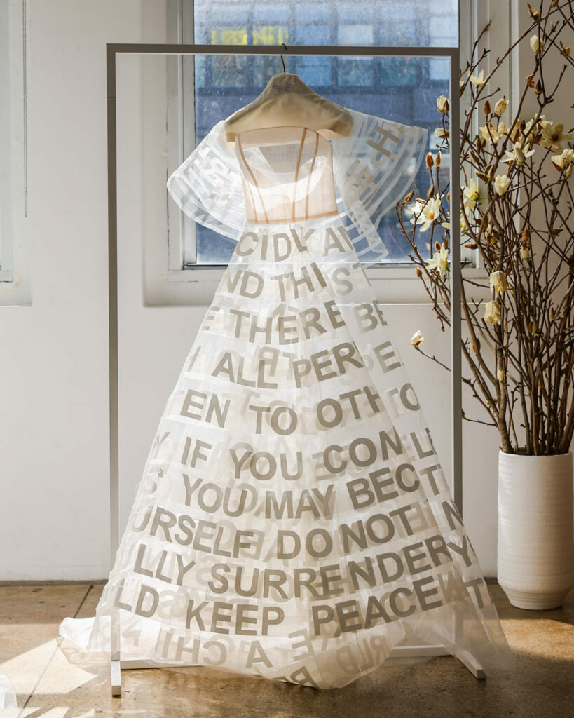 Sheer white SS2026 dress with bold gray letters in sunlit room.