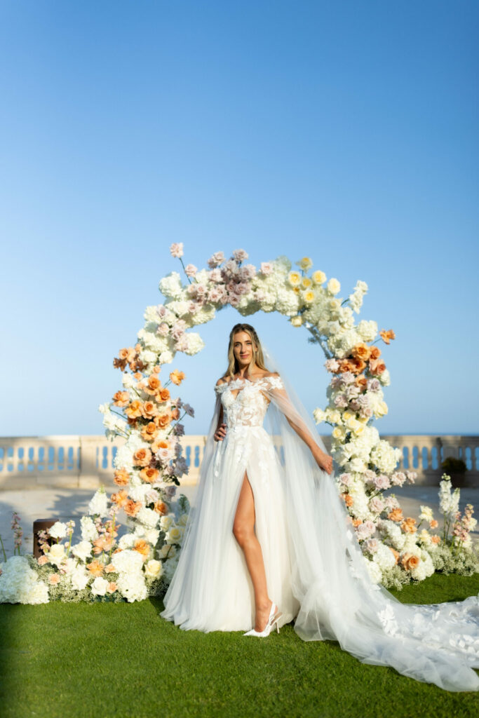 Bride before floral arch, blue sky, stone railing.