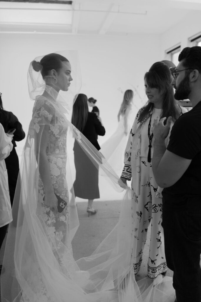 Bride in lace gown, veil adjusted by team, busy room.