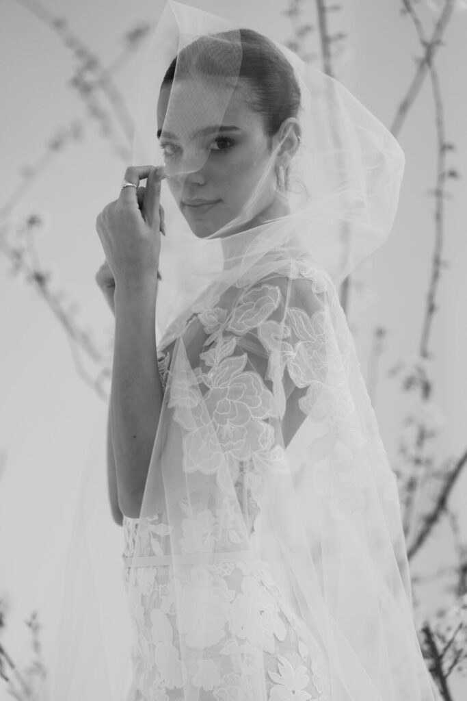 Woman in lace wedding dress, sheer veil, outdoors.