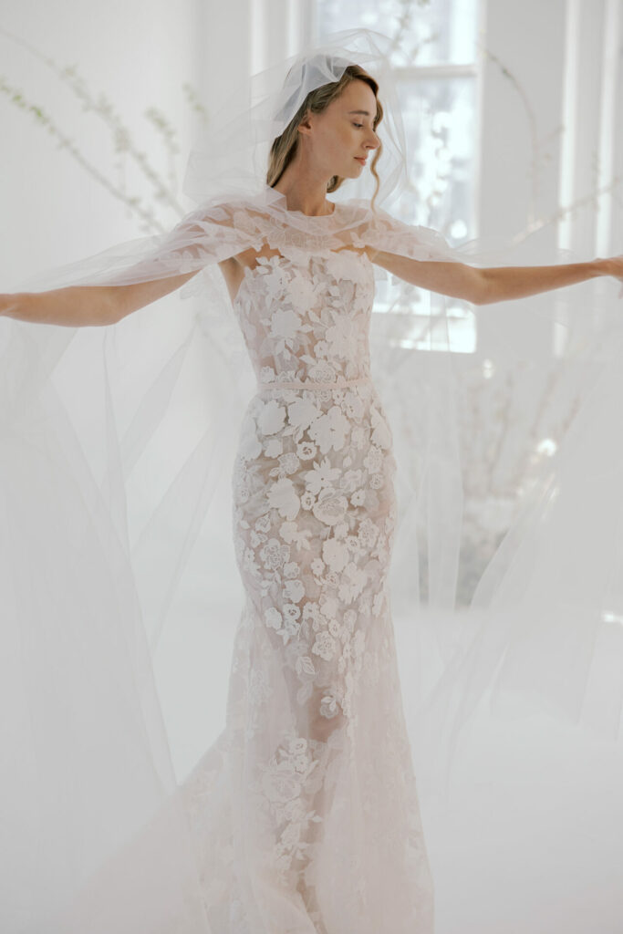 Bride in lace gown with veil, arms outstretched.