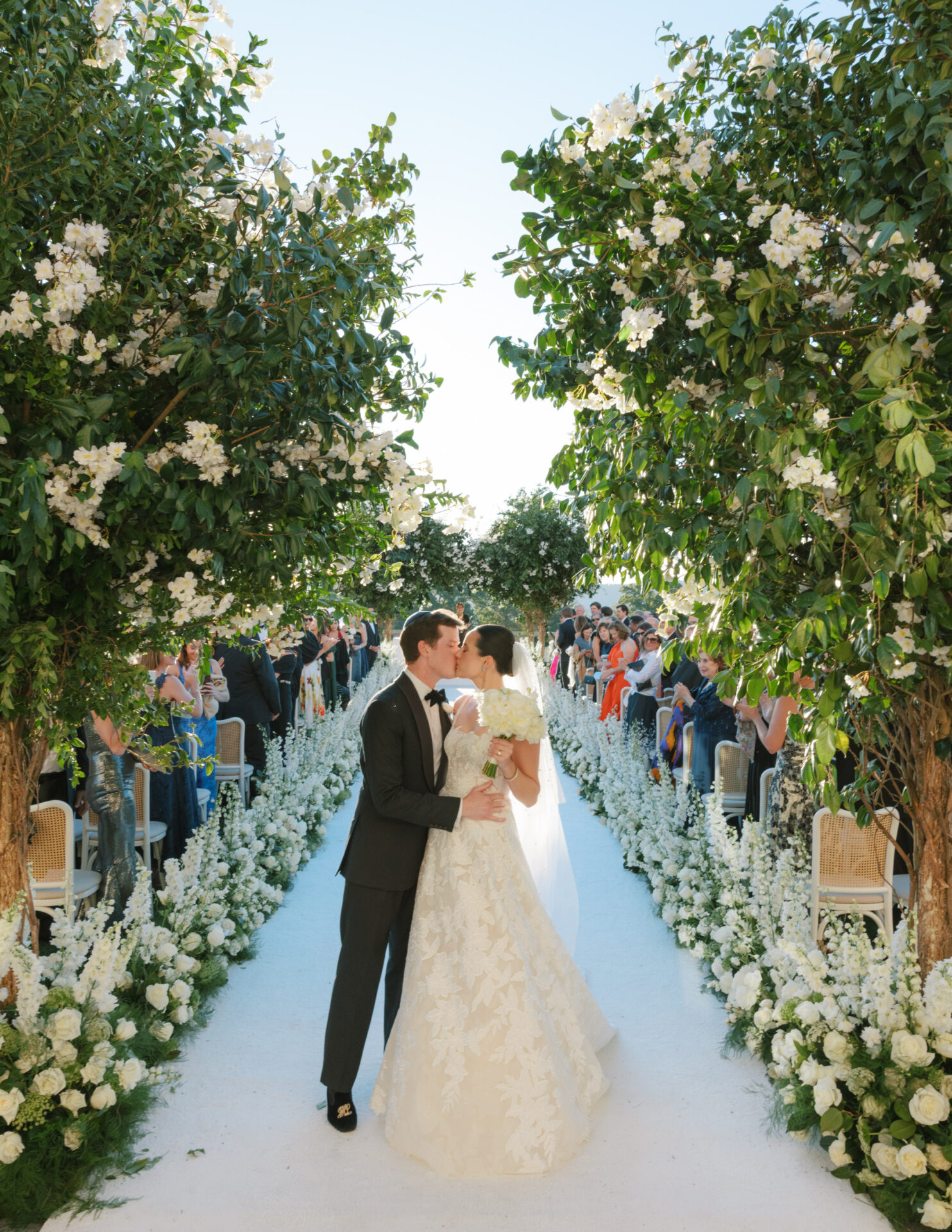 Bride and groom kiss in flower-lined outdoor aisle.