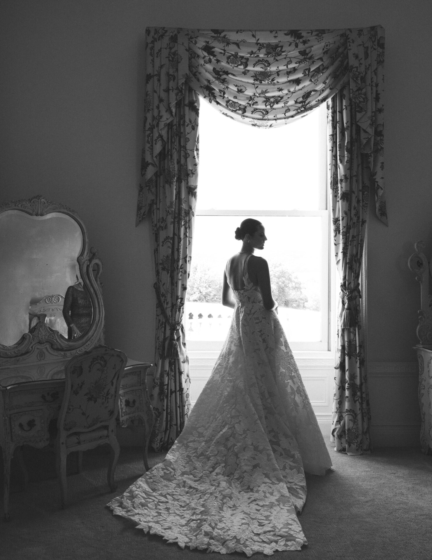 Bride in elegant gown gazes out ornate window.