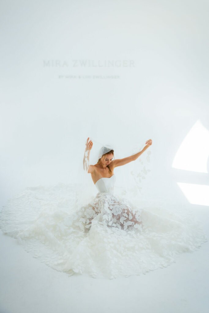 Bride in white gown holds floral veil, ethereal scene.