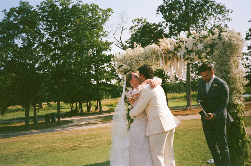 Bride and groom kiss under floral arch, man nearby.