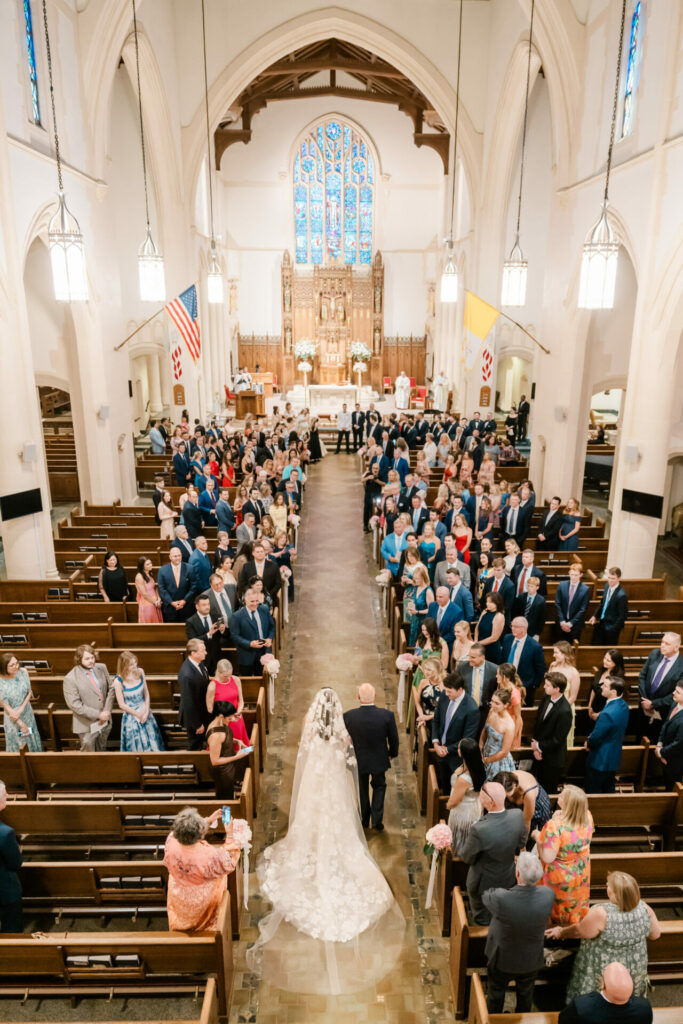 Bride in white gown walks aisle of ornate church.