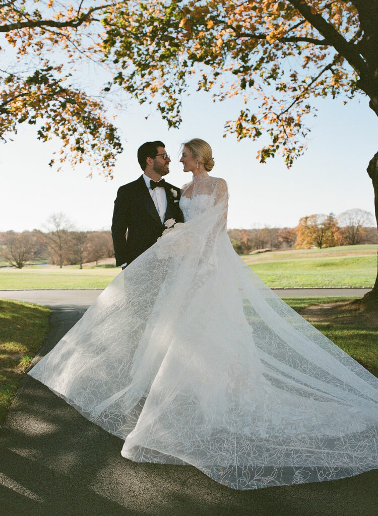 Bride and groom smile under autumn tree outdoors.