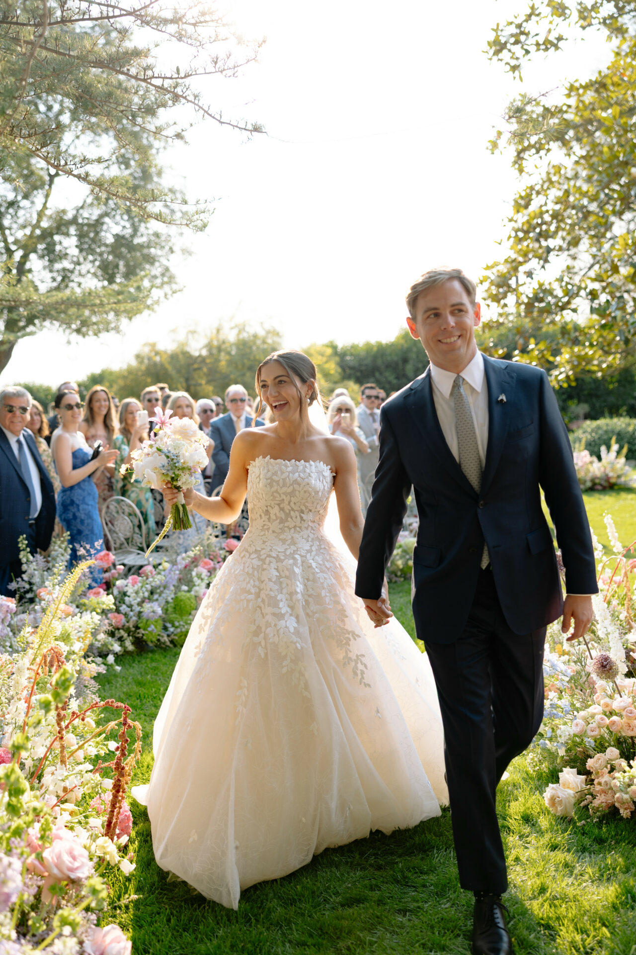 Bride and groom walk outdoors amid flowers, smiling.