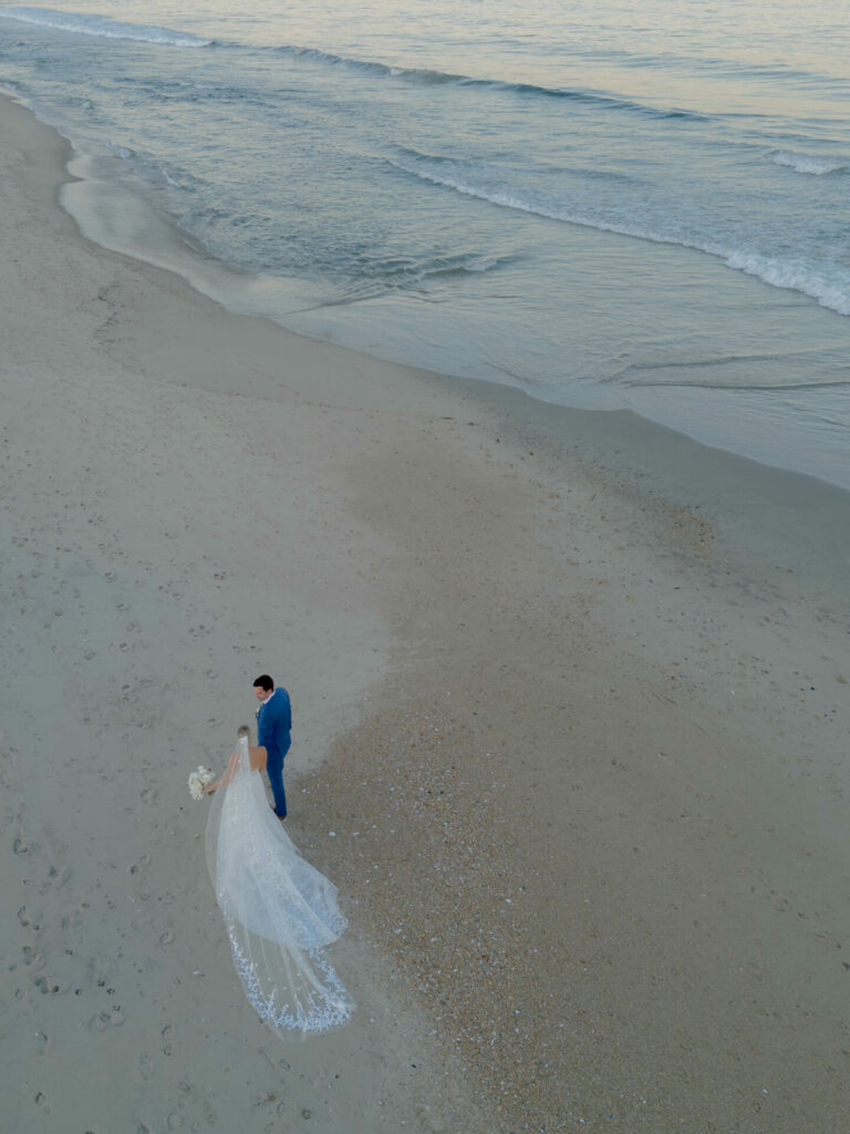 Bride and groom embrace on serene beach, flowing veil.