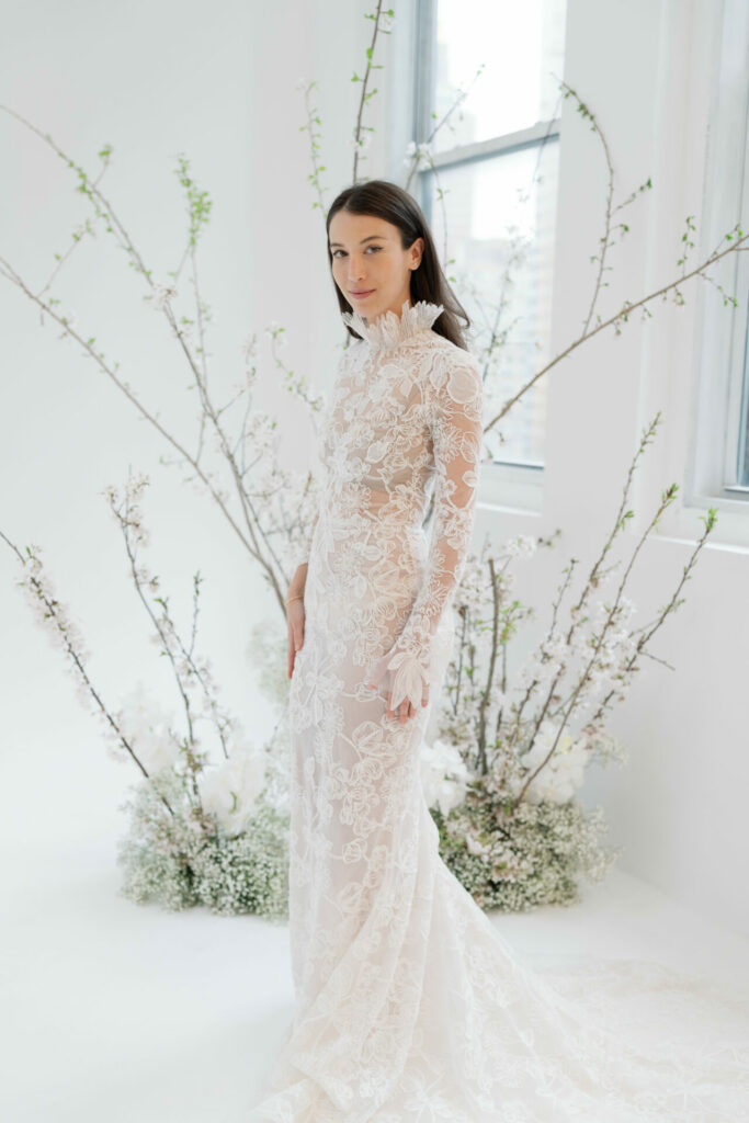 Bride in lace gown stands by window, florals.
