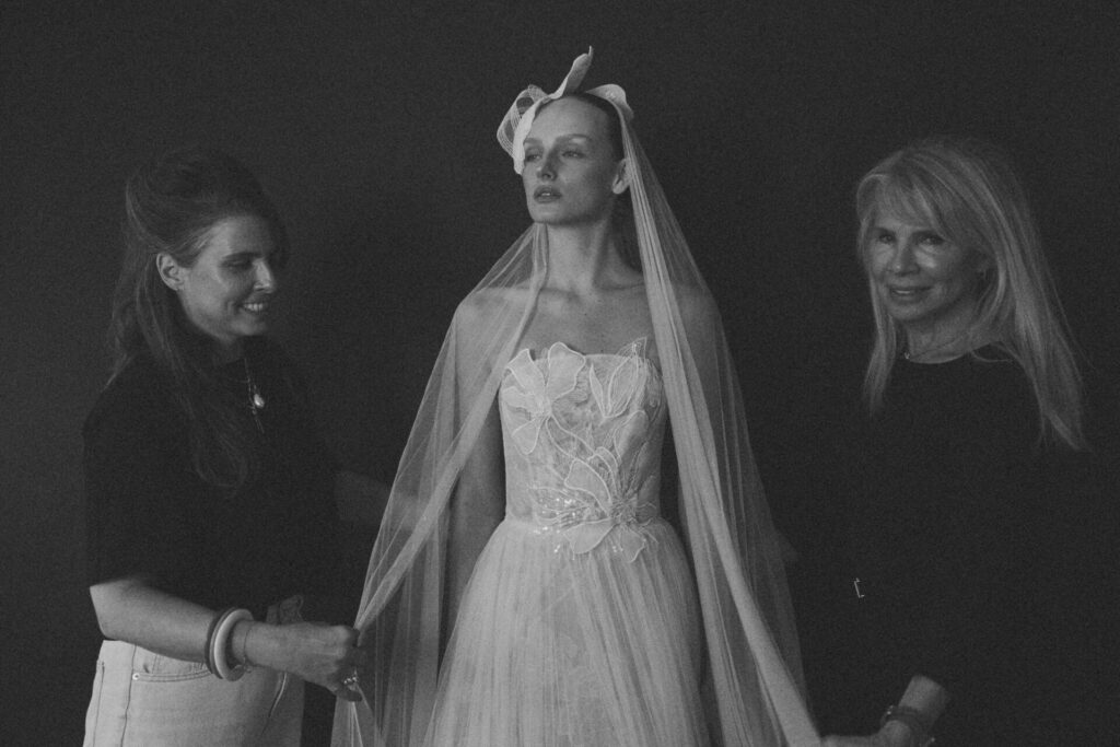 Bride in strapless gown, veil; two women adjusting.