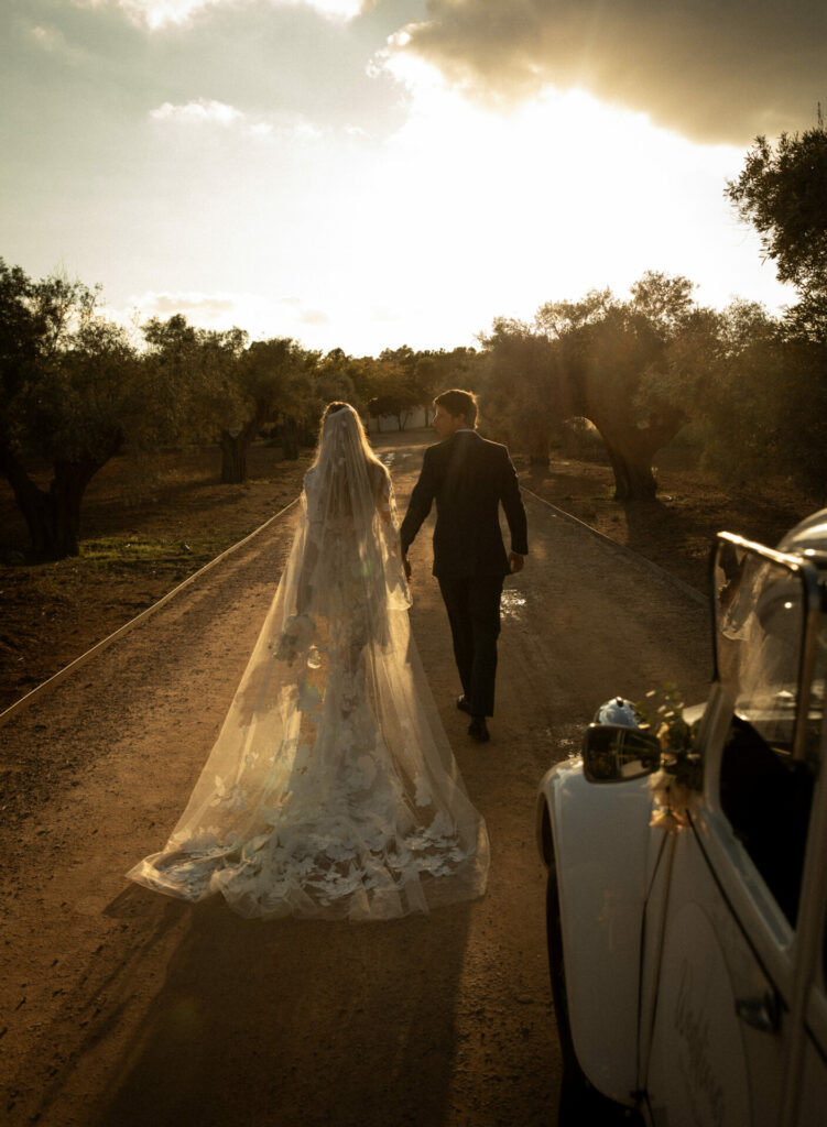 Bride and groom walk at sunset, vintage car nearby.